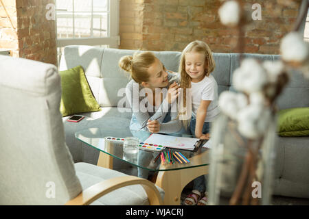 Female caucasian teacher and little girl, or mom and daughter. Homeschooling. Sitting on the sofa and drawing with paints, writing, talking and having fun. Education, school, studying concept. Stock Photo