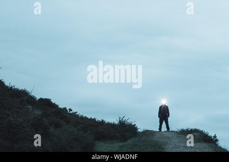 A scary silhoutted figure standing on the top of a path on a hill in the countryside, with a light glowing from his head. On a winters evening. Stock Photo