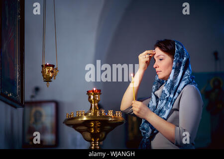 Russian beautiful Caucasian woman with red hair and a scarf on her head is in the Orthodox Church, lights a candle and prays in front of the icon Stock Photo