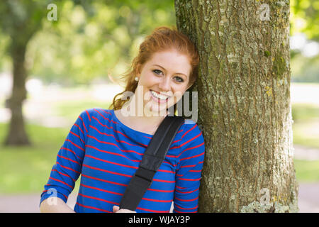 Happy redhead student leaning on tree looking at camera Stock Photo