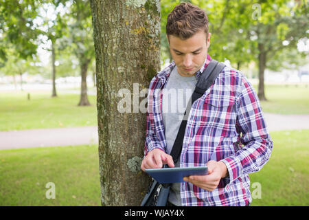 Handsome student leaning on tree using his tablet pc Stock Photo