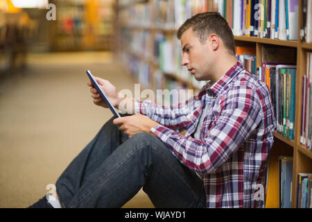 Thoughtful young student sitting on library floor using tablet Stock Photo