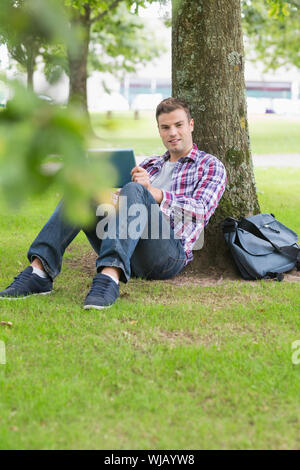 Smiling student using his tablet pc outside leaning on tree Stock Photo