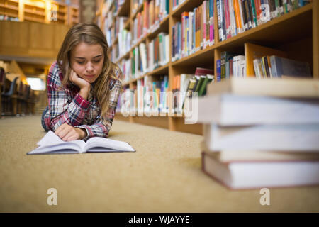 Pretty focused student lying on library floor reading book Stock Photo