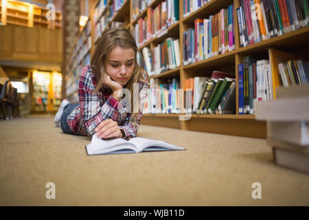 Pretty focused student lying on library floor reading book Stock Photo