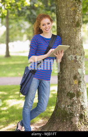 Smiling student leaning against a tree using her tablet pc Stock Photo