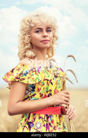 Portrait of a girl with curly blond hair, holding ripe spikelets of barley in her hands. A model in a colored sundress poses on the background of an a Stock Photo