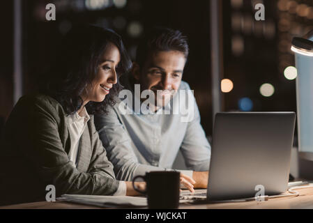 Two smiling office colleagues using a laptop while working overtime Stock Photo