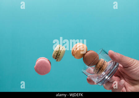 A hand holding a glass tumbler with five coloured macaroons floating on a blue background. Pink, brown, green, yellow and beige Macaroons. Stock Photo