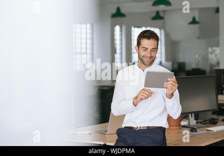 Smiling businessman leaning on an office desk using a tablet Stock Photo