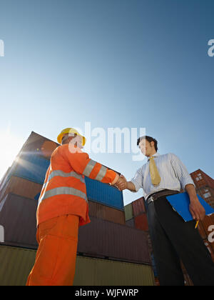 Mid adult businessman holding clipboard and shaking hands to manual worker near cargo containers. Vertical shape, low angle view. Copy space Stock Photo