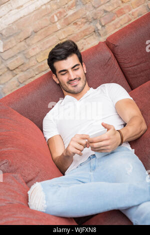 A smiling handsome young man in a white tshirt laying on a sofa and using his smartphone. Stock Photo