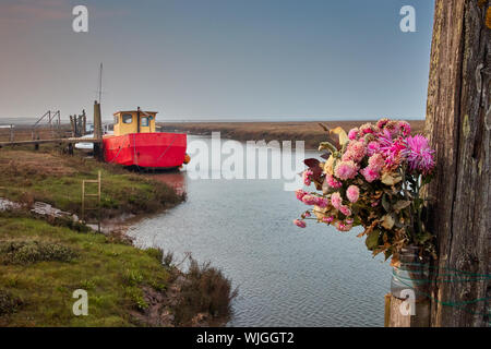 A bouquet of pink and lilac flowers tied to a post at the side of a saltmarsh creek near Thornham, Norfolk with a fishing boat in the background Stock Photo