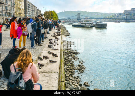 Tourists visit and take photos at The Shoes on the Danube Bank, a memorial in Budapest, Hungary with the Chain Bridge and boats in view Stock Photo