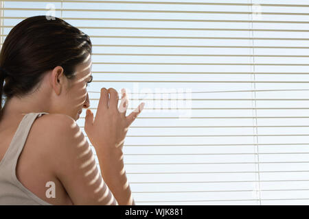 Rear view of young woman peering through blinds at home Stock Photo