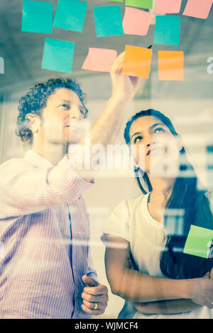 Businessman and businesswoman discussing over adhesive notes on glass wall in office Stock Photo