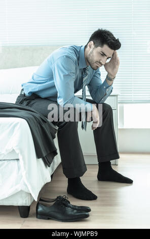 Wincing businessman sitting at edge of bed at home in bedroom Stock Photo