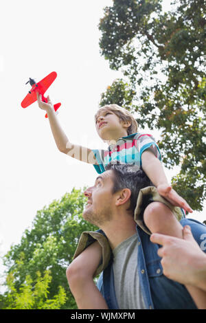 Low angle view of a boy with toy aeroplane sitting on father's shoulders at the park Stock Photo