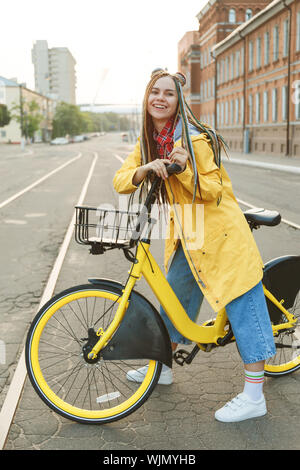 Young woman wearing yellow coat and colored pigtails, riding bike in city. Stock Photo