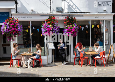 Winchester, Hampshire, UK the front of a cafe with people sitting at tables in the sun enjoying food and a coffee with a waitress carrying Stock Photo