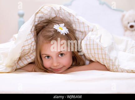 Sweet little girl lying down in the bed covered with blanket, cute flower in hair, sleeping in child bedroom, happy childhood concept Stock Photo