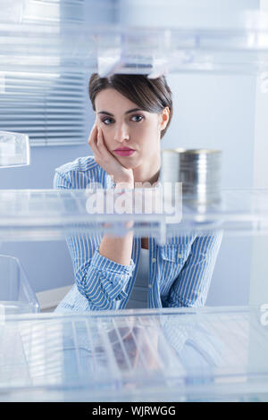 Young sad woman looking at one tin in her empty fridge. Stock Photo