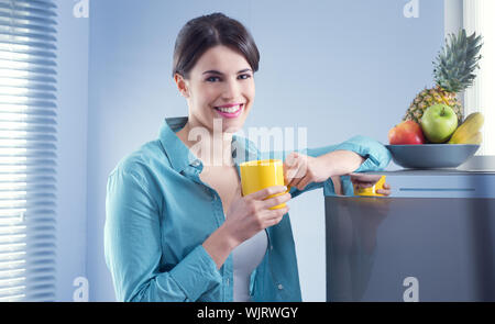 Young cheerful woman holding a yellow mug and leaning against refrigerator. Stock Photo