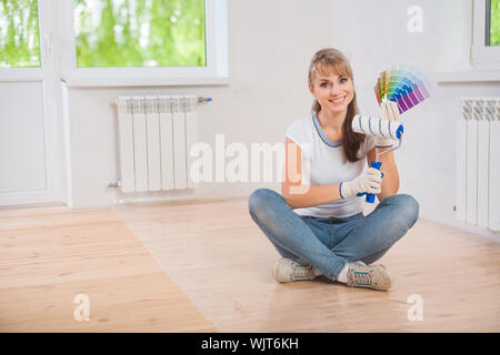 beautiful smiling female worker sitting on wooden floor and showing paintroller and color palette Stock Photo