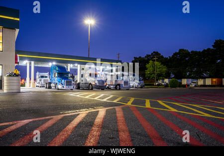 Different make and models big rigs semi trucks with semi trailers standing on the truck stop parking lot under the lighted shelter in night and comply Stock Photo