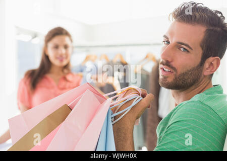 Bored man sitting with shopping bags while woman by clothes rack in the background Stock Photo