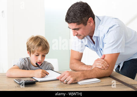 Son doing homework while father standing by at table in house Stock Photo