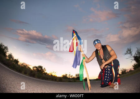 Indigenous man kneeling in the middle of a road with ceremonial pole representing seven directions Stock Photo