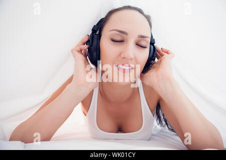Happy brunette lying under the sheets listening to music Stock Photo
