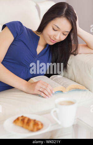 Pretty young asian woman lying on the sofa reading a book Stock Photo