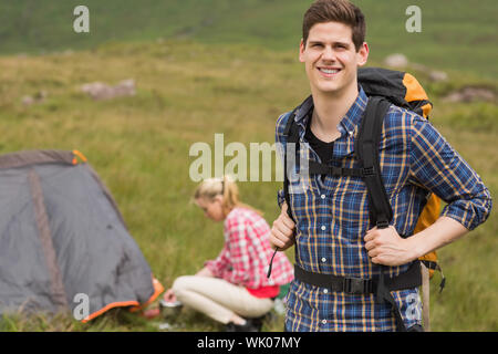 Cheerful man carrying backpack while girlfriend is pitching tent Stock Photo