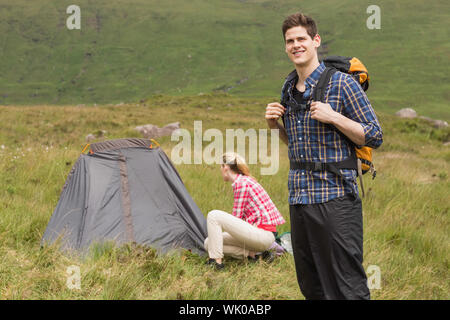 Smiling man carrying backpack while girlfriend is pitching tent Stock Photo
