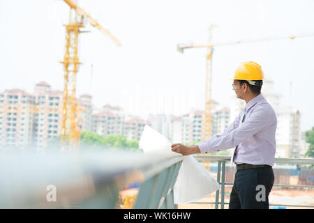 Portrait of a smiling Asian Indian male contractor engineer with hard hat inspecting at construction site. Stock Photo