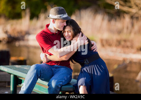 Young Caucasian man hugging his biracial girlfriend, sitting together on dock over lake Stock Photo