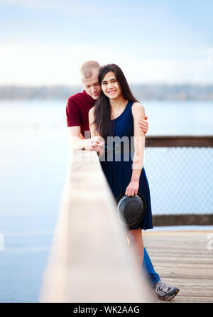 Young happy interracial couple standing together on wooden pier overlooking lake. Man bowing head in prayer. Stock Photo