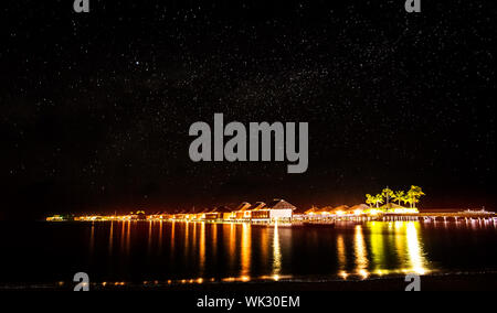 Night on tropical resort, many little beach houses glowing bright yellow lights in dark night, starry sky over sea, romantic summer holidays concept Stock Photo