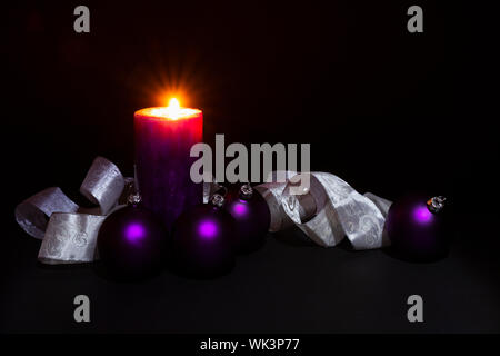Arrangement of beautiful illuminated purple colored Christmas balls with a silver and white colored ornate ribbon and a burning candle on black backgr Stock Photo
