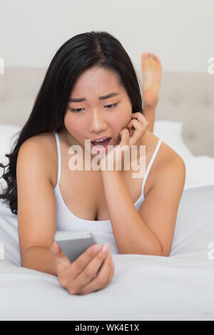 Shocked woman lying on bed holding smartphone at home in bedroom Stock Photo