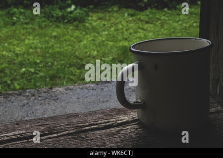 Very old with a lot of scratches white metallic mug in front of the green grass background and placed on wooden plank with copy space Stock Photo