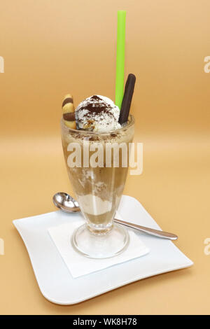 Iced coffee float topped with vanilla ice cream, cocoa powder, chocolate stick, and wafer roll, served in a tall glass with green straw and spoon. Stock Photo