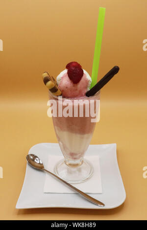 Tall glass of strawberry and cream milkshake topped with raspberry and wafer sticks, served with a green straw and a spoon on a white plate Stock Photo