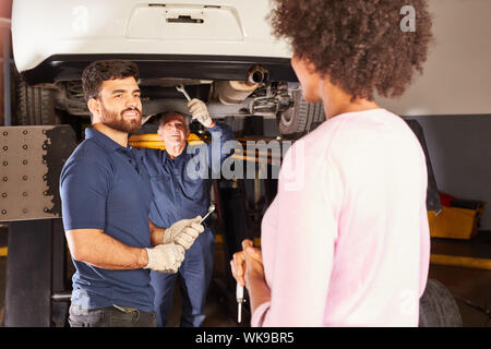 Two car mechanics repair car on lift with customer in foreground Stock Photo