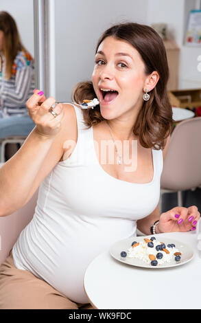 Portrait of young pregnant woman wearing white shirt eating cottage cheese with berries and nuts in a cafe - healthy nutrition and pregnancy concept Stock Photo