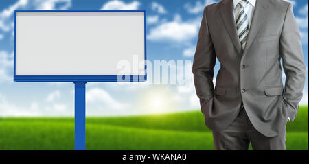 Businessman standing with hands in pockets. Blank billboard, blue sky and green grass as backdrop Stock Photo