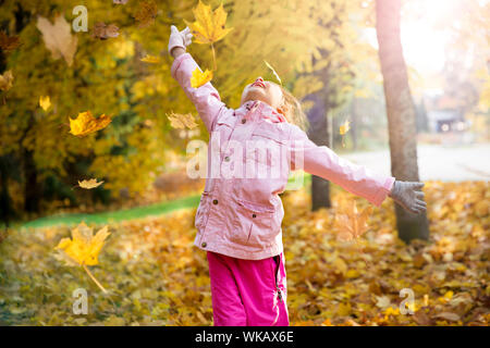 Cute little girl with missing teeth playing with yellow fallen leaves in autumn forest, trowing into the air. Happy child laughing and smiling. Sunny Stock Photo