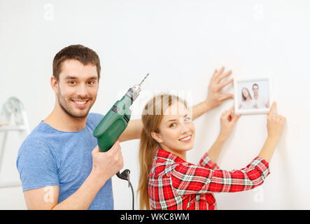 repair, interior design, building, renovation and home concept - smiling couple drilling hole in wall and putting picture up at home Stock Photo